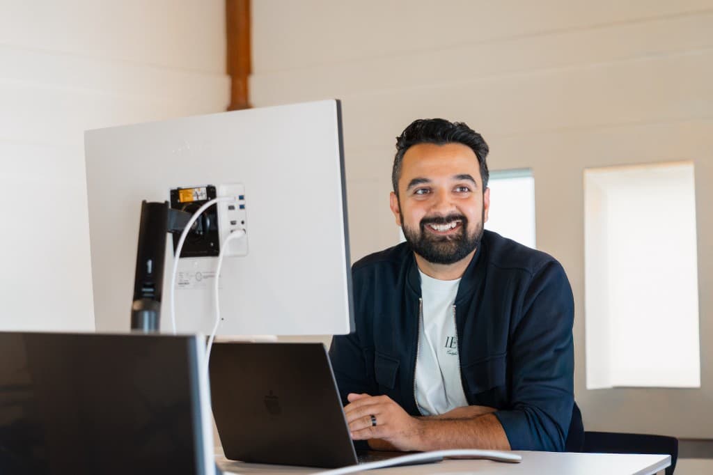 A Cohorvaer smiling at a desk beside a monitor and laptop