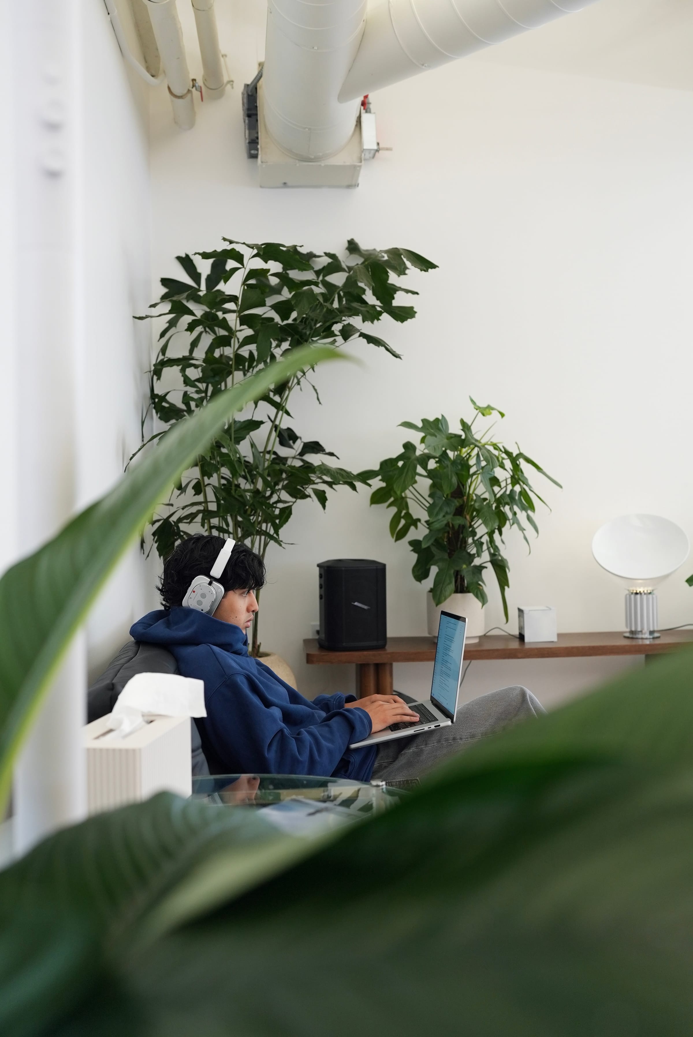 A Cohorvaer working from the office couch in headphones, framed by plants